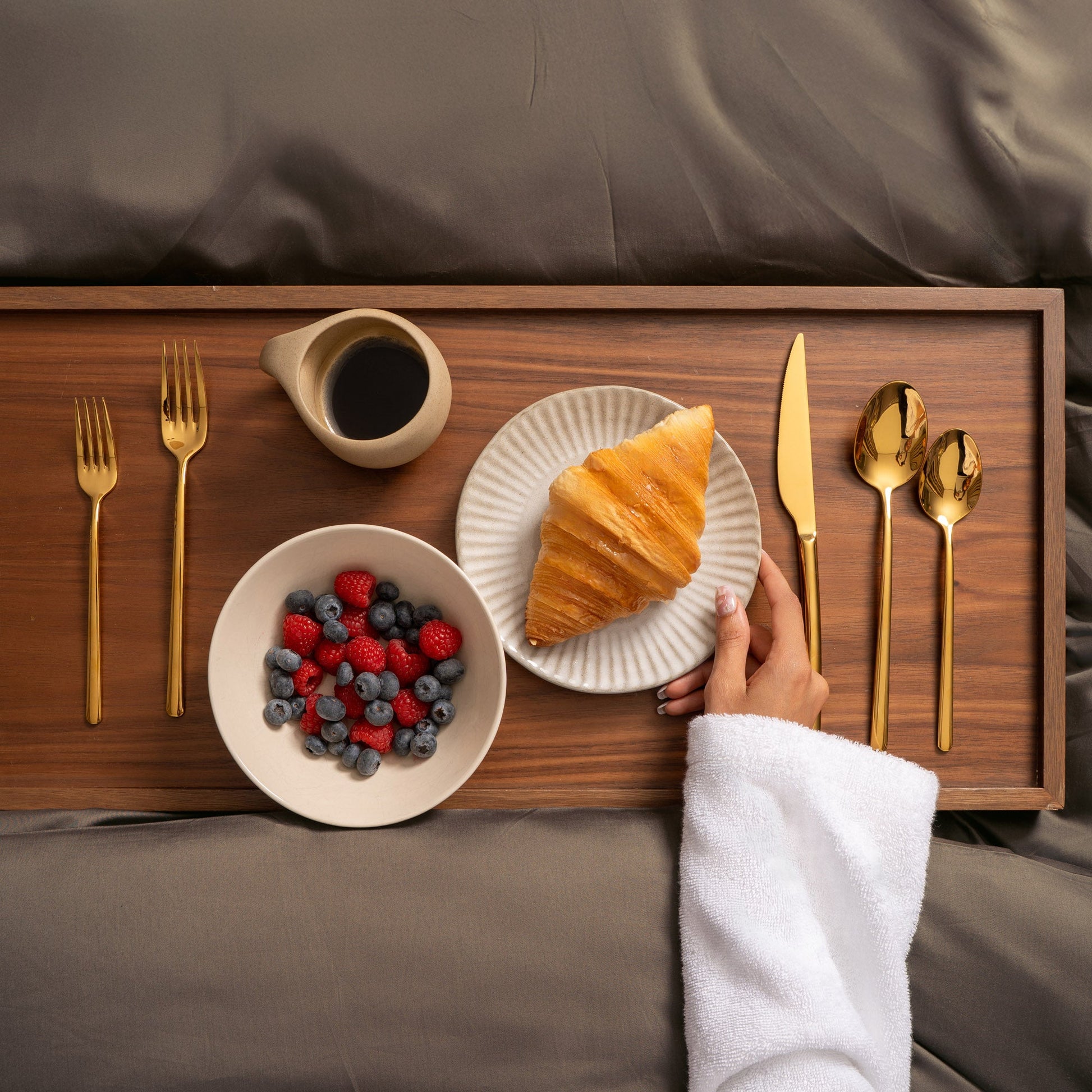 Wooden tray with breakfast items including a croissant, berries, coffee, and cutlery on a bed.