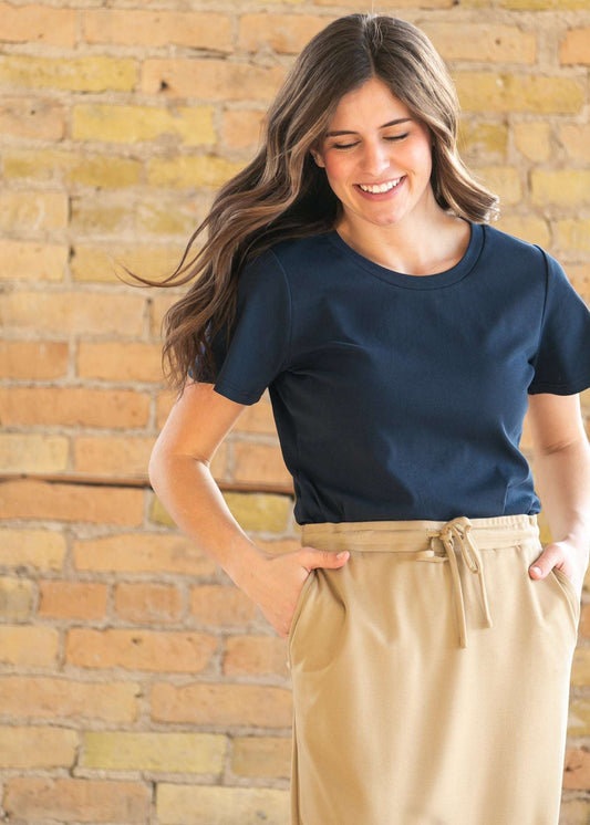 A woman smiling and posing in front of a brick wall, wearing a navy blue short-sleeve crewneck t-shirt and beige high-waisted drawstring trousers.