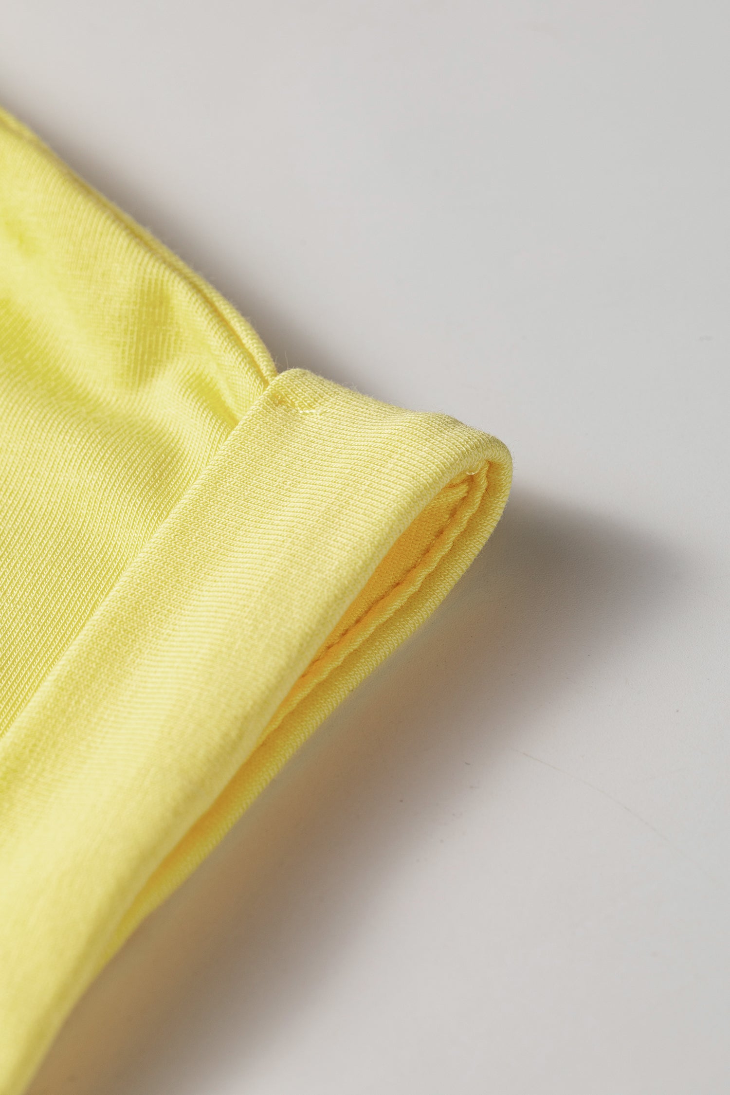 A close-up detail shot of a folded light yellow fabric item, showing the texture and stitching on a neutral light gray background.