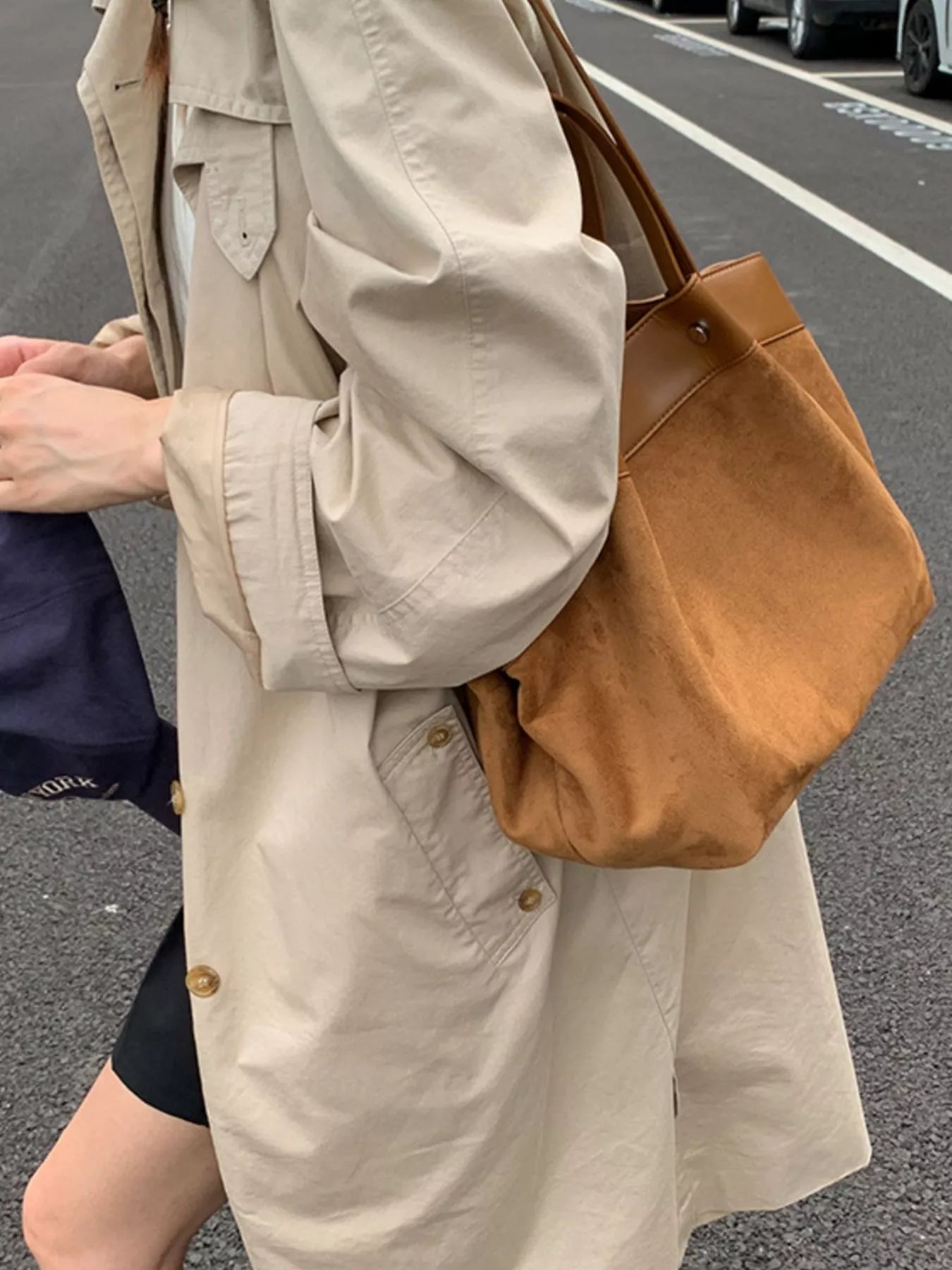A person wearing a beige trench coat carries a large brown suede or faux-suede shoulder bag on a city street.