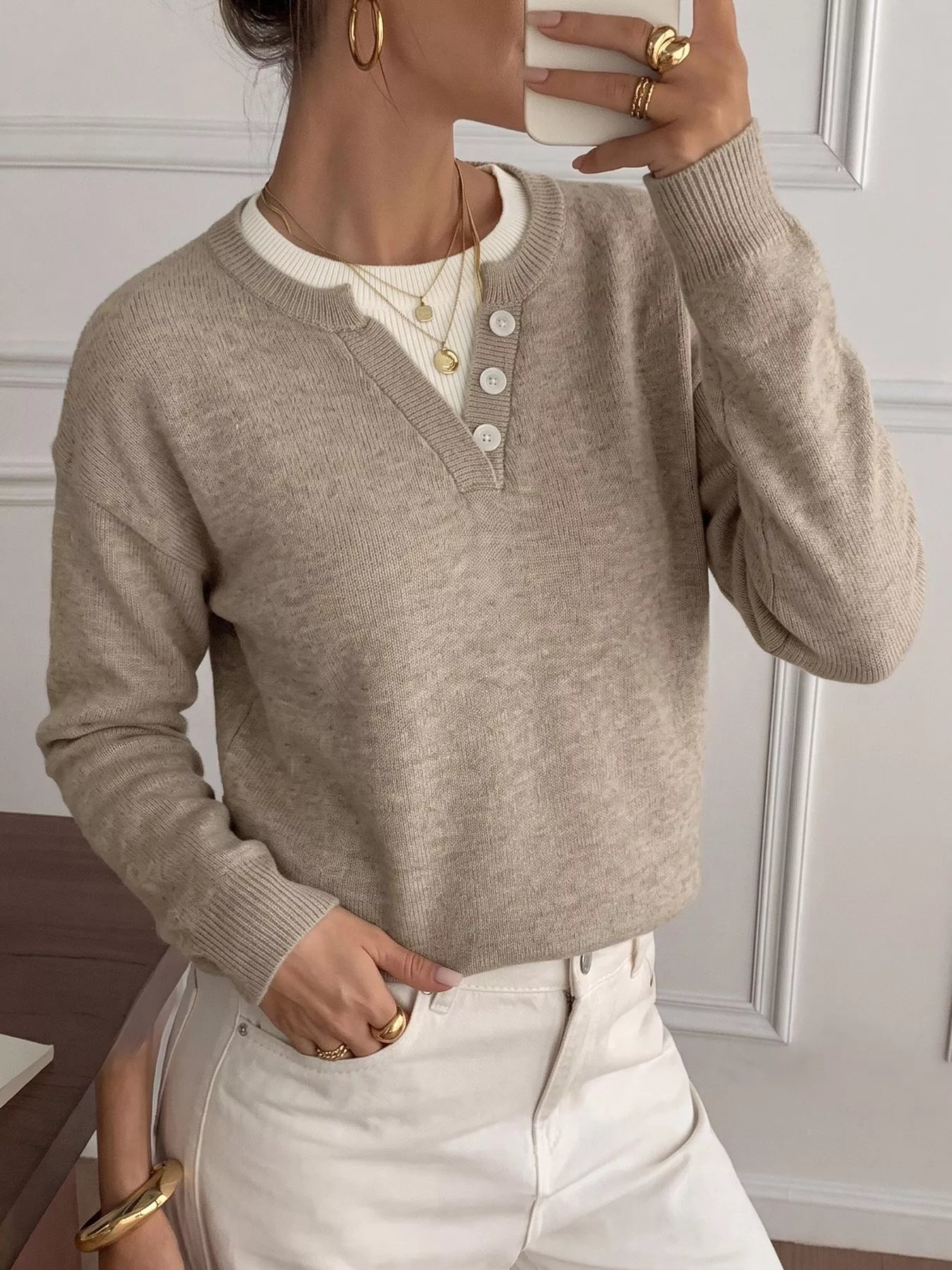 A woman wearing a light taupe knit sweater with a buttoned henley-style neckline over a cream shirt, paired with white pants. She is holding a white cup and wearing gold jewelry in an indoor setting with a paneled wall background.