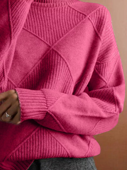 A close-up of a person wearing a chunky knit sweater in a vibrant magenta pink. The sweater features a diamond-shaped geometric pattern and ribbed textures on the sleeves and shoulders, paired with gray trousers.