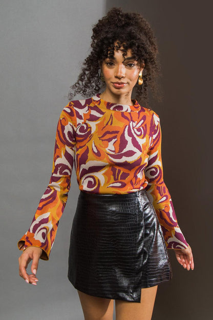 Woman wearing a patterned orange and purple top with a black leather skirt against a gray background