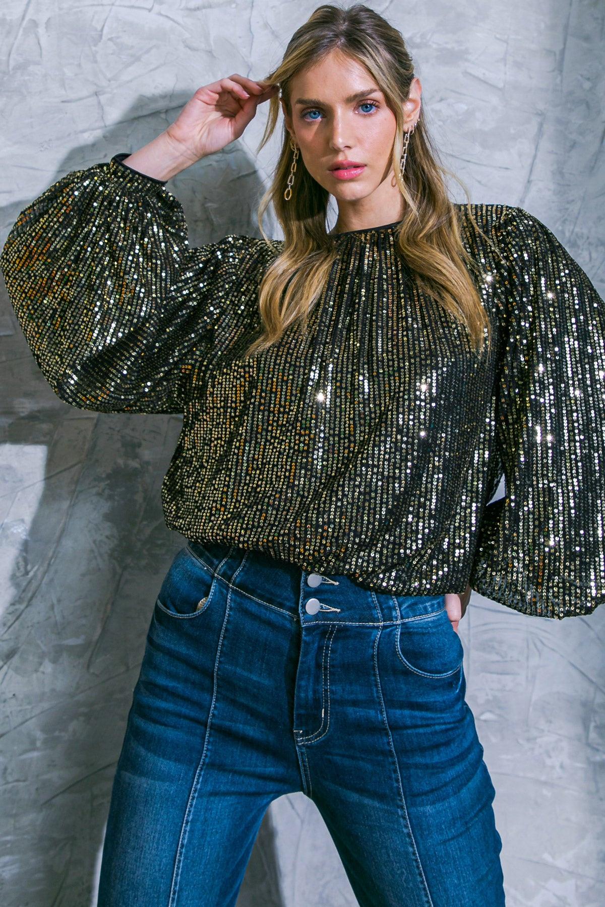 Woman wearing a sparkling top and blue jeans against a textured gray wall.