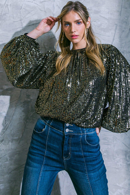 Woman wearing a sparkling top and blue jeans against a textured gray wall.