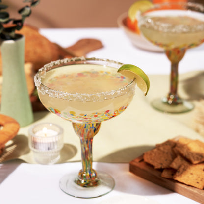 Colorful speckled margarita glass with a salted rim and lime wedge on a table beside tortilla chips.
