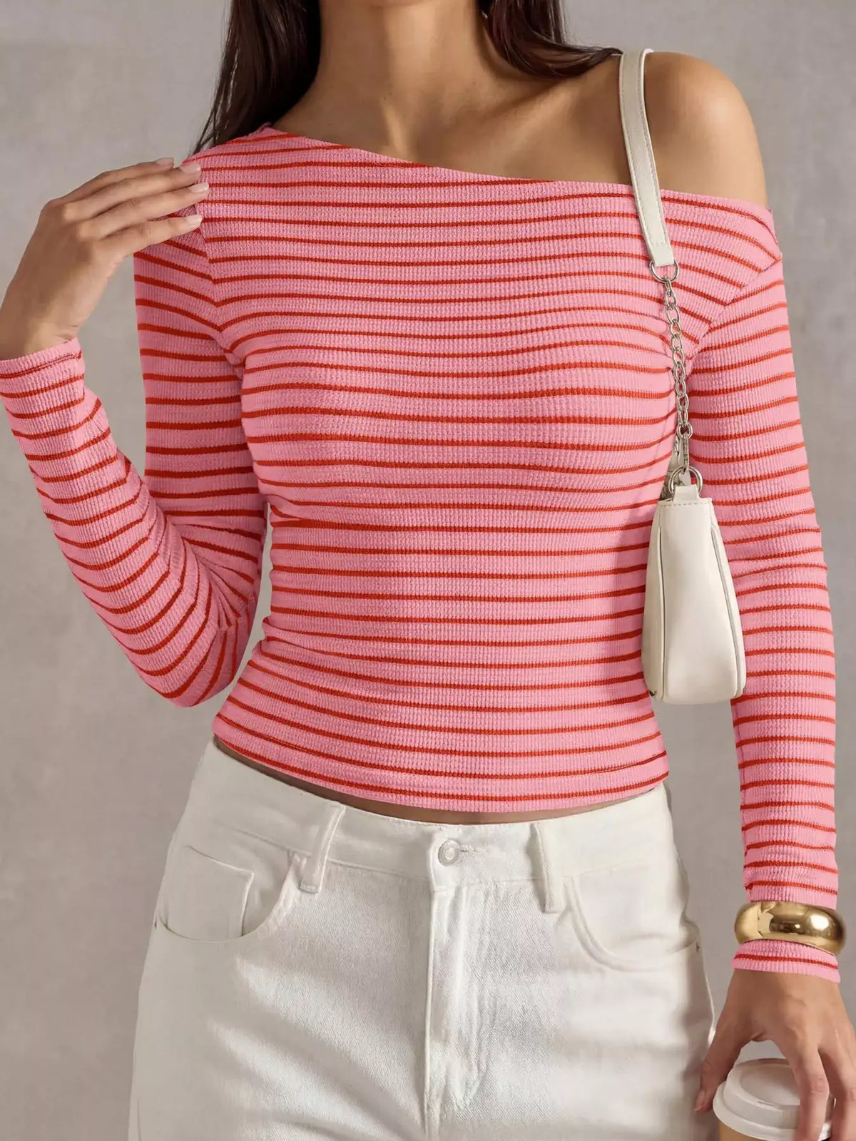A woman wearing a coral pink and red horizontally striped off-the-shoulder long-sleeve ribbed top, paired with white pants and a small cream shoulder bag.