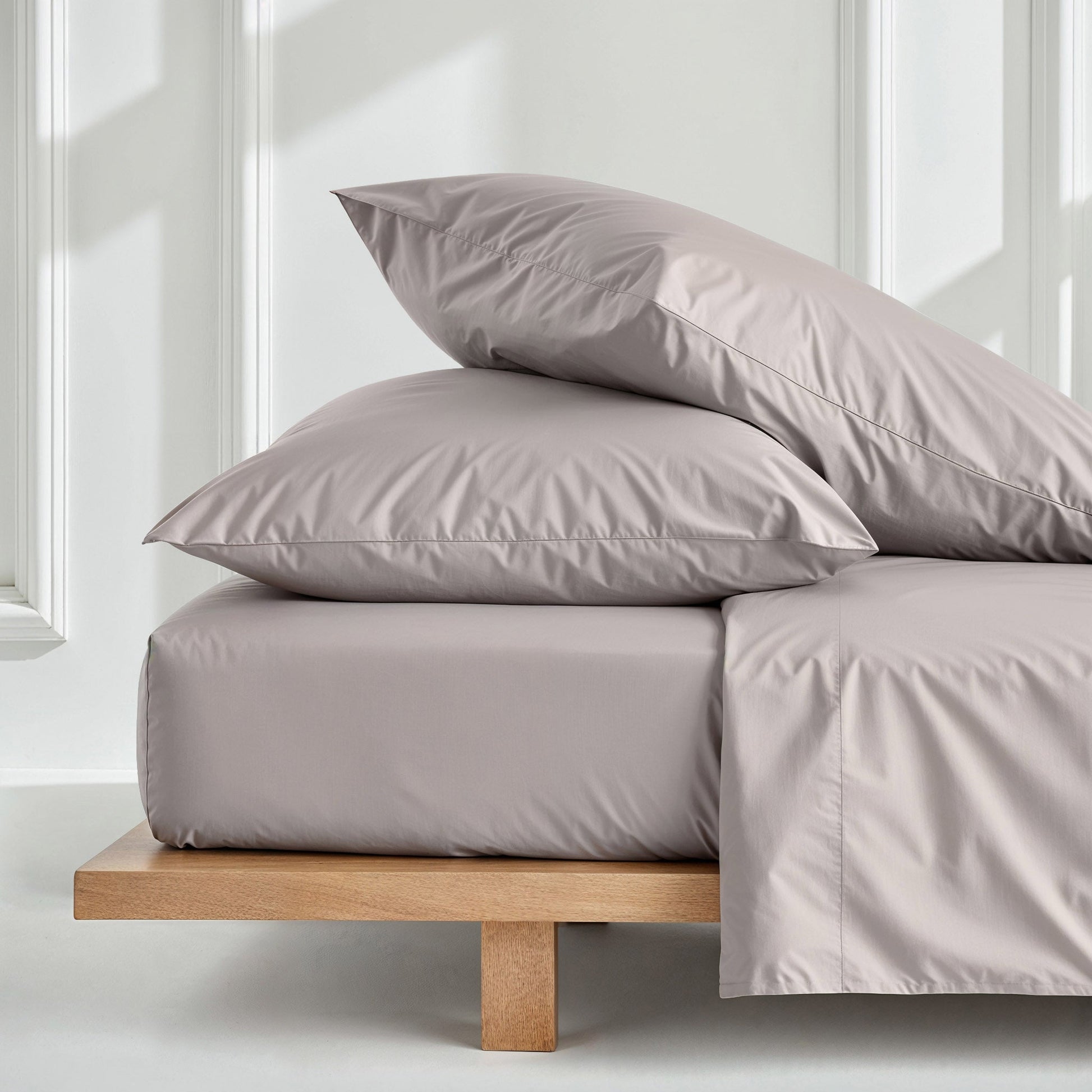 A bed dressed in a light taupe-gray bedding set with two matching pillows and a folded sheet, resting on a low wooden platform bed frame in a bright white room.