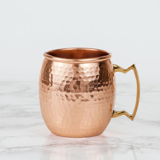 Hammered copper Moscow mule mug with a gold-tone handle on a white marble surface.