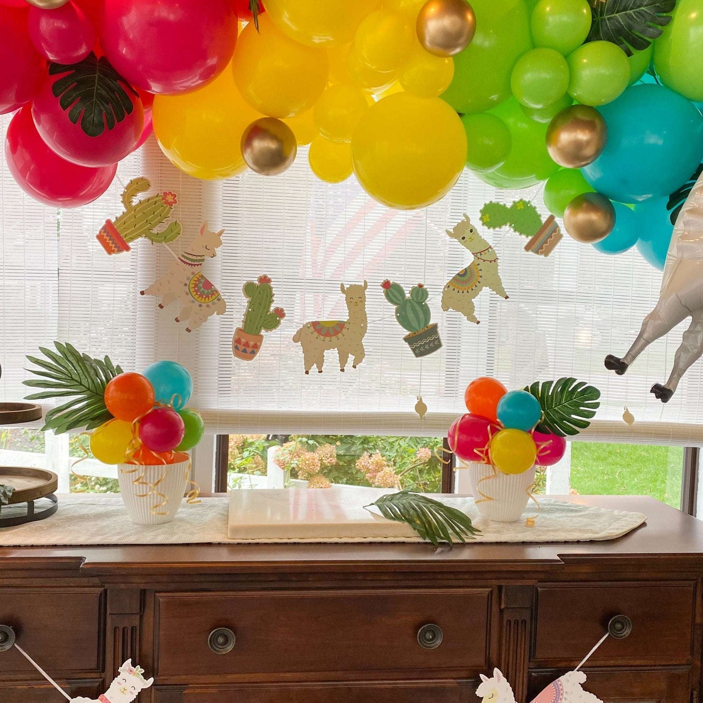 Colorful balloon garland with llama and cactus cutouts hanging over a wooden dresser with two small balloon centerpieces.