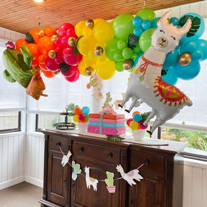 Rainbow balloon garland with llama and cactus foil balloons, tropical leaves, and a llama banner on a wood cabinet.