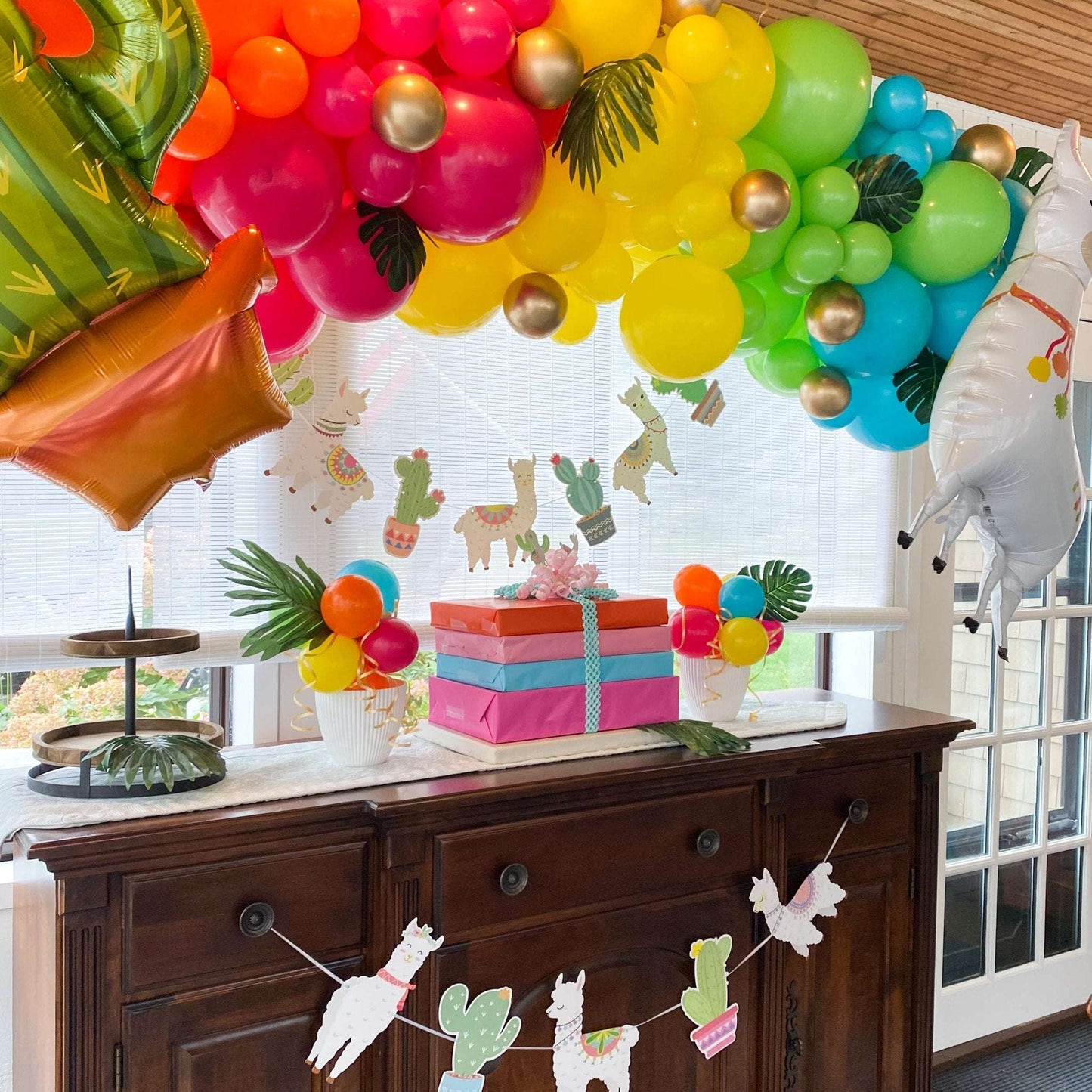 Rainbow balloon garland with gold accents, llama and cactus foil balloons, and paper banners over a wooden sideboard.