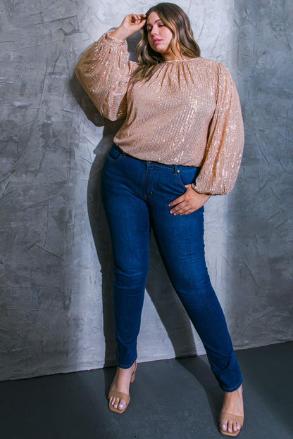 Woman wearing a sparkly top and blue jeans against a gray wall.