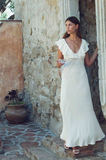 A woman wearing a white ruffled off-the-shoulder maxi dress stands in a rustic stone doorway with a potted plant nearby.