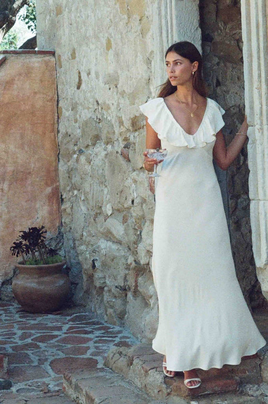 A woman wearing a white ruffled off-the-shoulder maxi dress stands in a rustic stone doorway with a potted plant nearby.