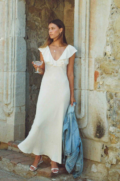 Woman in a white dress holding a blue jacket and a glass, standing against a stone wall.