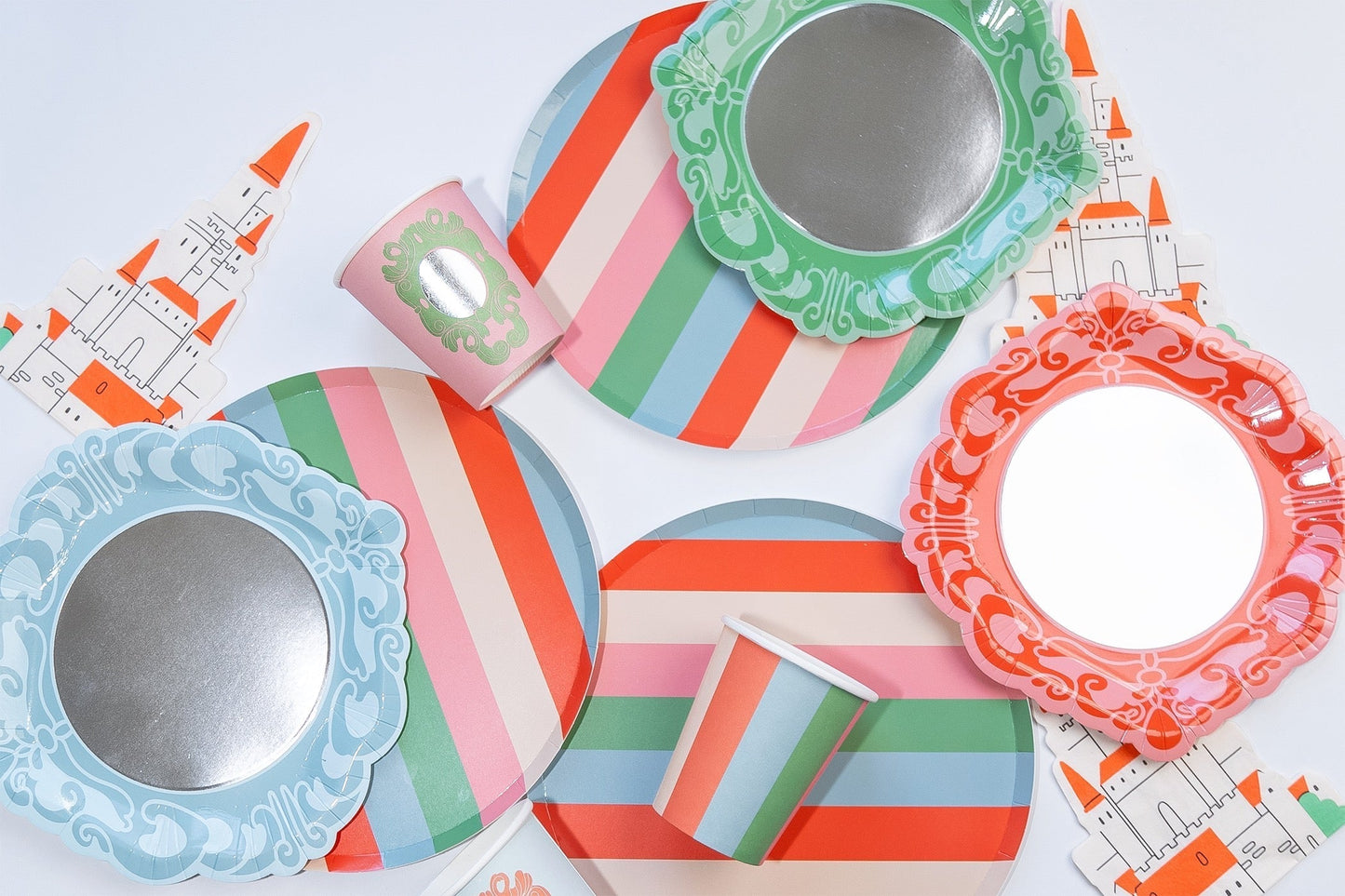 Striped paper plates, cups, and mirror-shaped plates with castle cutouts in pink, blue, and green on a white surface.