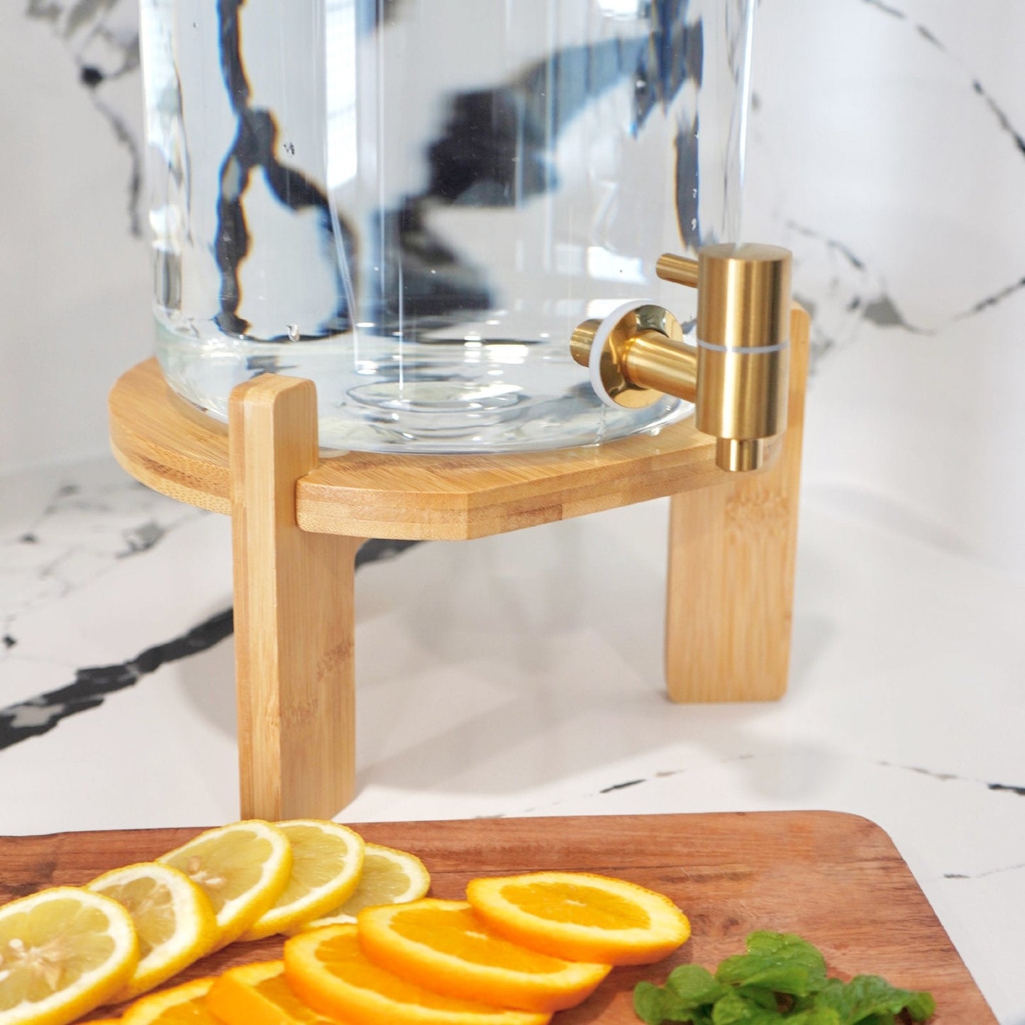 Clear glass carafe with gold accents on a wooden stand, surrounded by sliced lemons and oranges on a marble surface.