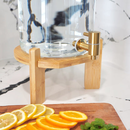 Clear glass carafe with gold accents on a wooden stand, surrounded by sliced lemons and oranges on a marble surface.