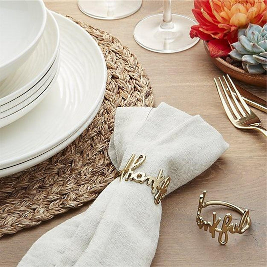 Table setting with white plates, gold 'thankful' napkin rings, and a woven placemat.