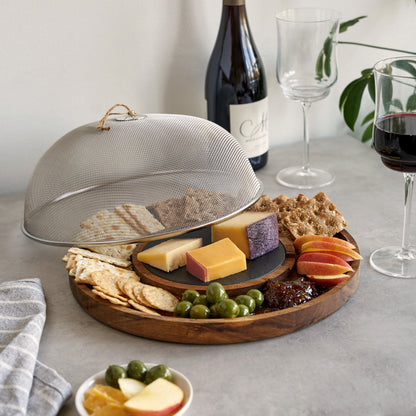 Wooden charcuterie board with assorted cheeses, fruits, and crackers on a table with a wine bottle and glasses.
