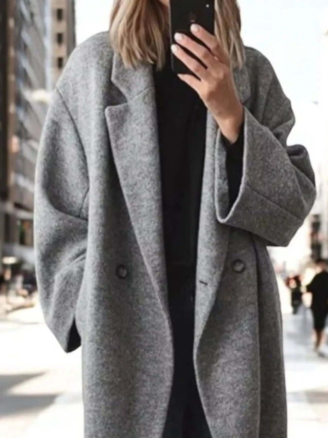 A person taking a mirror selfie wearing an oversized, double-breasted gray wool-blend coat over a black top, set against a blurred city street background.
