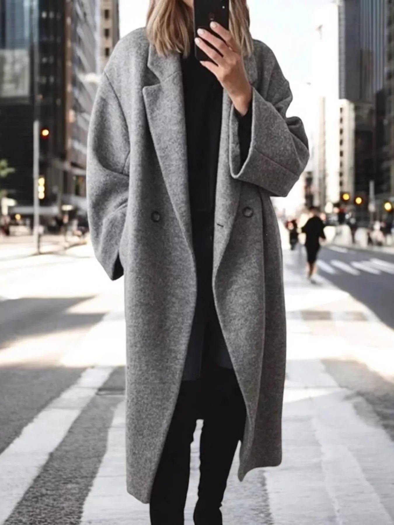 A woman takes a mirror selfie on a city crosswalk wearing a long, oversized gray wool-blend coat over a black outfit.
