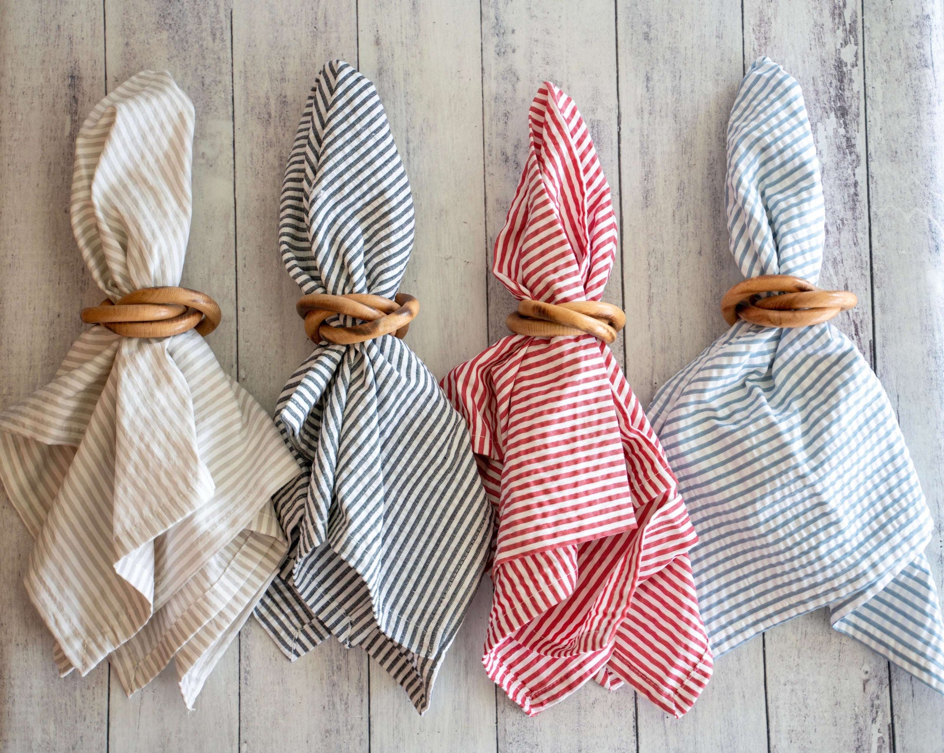 Four round wood napkin rings holding striped fabric napkins in tan, black, red, and blue on a light wood surface.