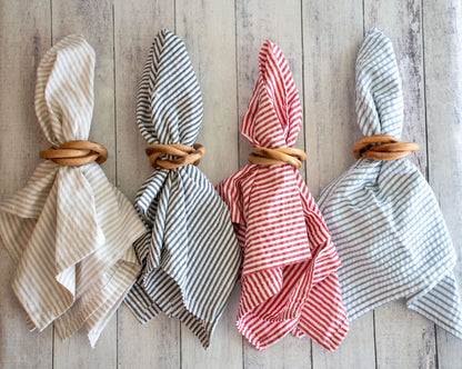 Four round wood napkin rings holding striped fabric napkins in tan, black, red, and blue on a light wood surface.