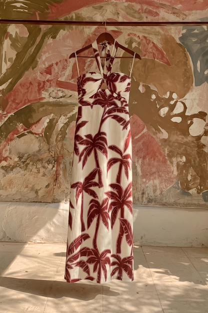 A sleeveless halter maxi dress with a red palm tree pattern on a white background, hanging on a wooden hanger against a textured, abstract painted wall.
