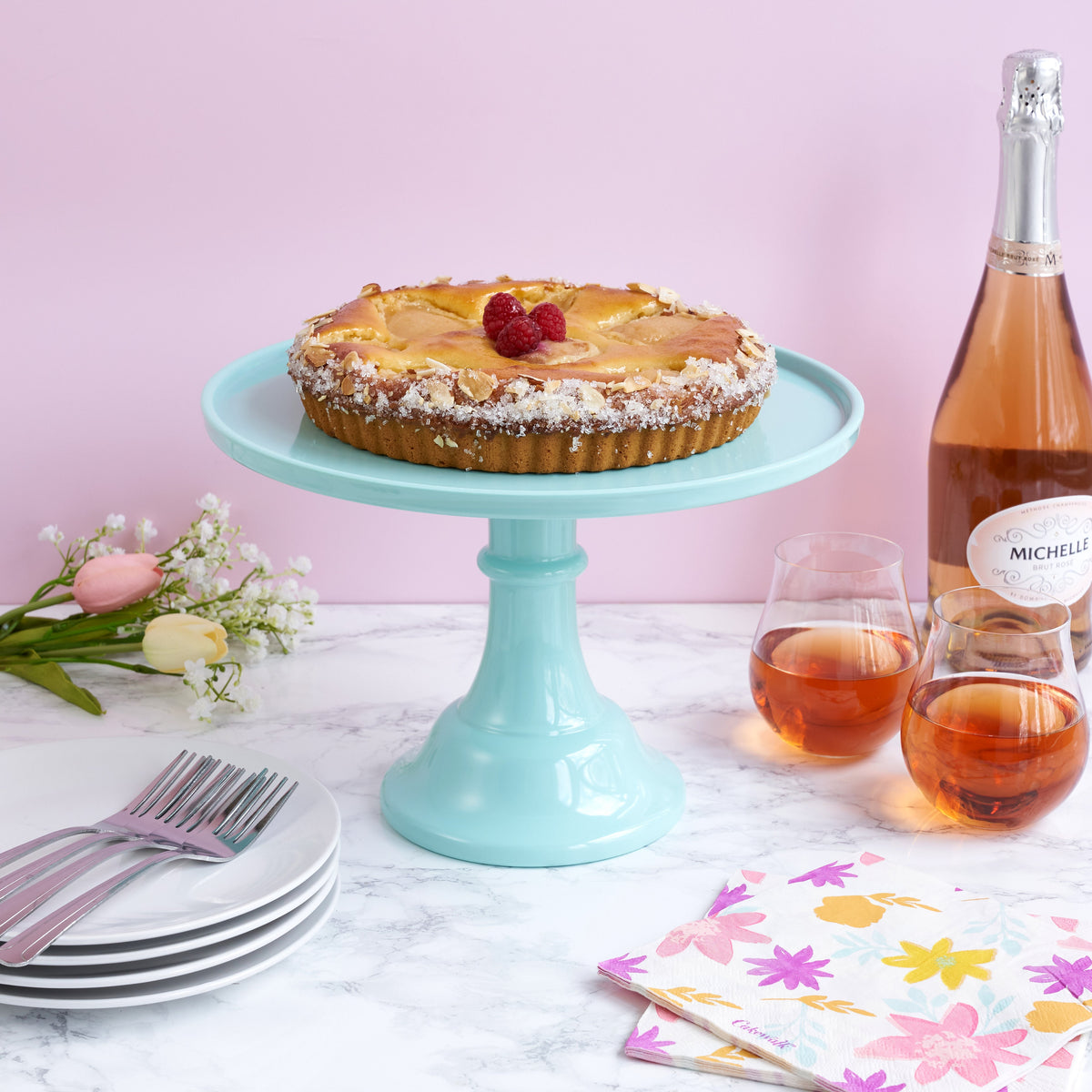 Melamine cake stand with a cake on it displayed on a table with wine, plates, napkins.