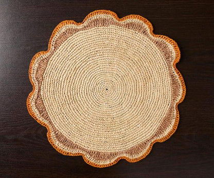 Round raffia woven placemat with a scalloped edge, featuring natural fibers in beige and orange tones on a dark wood surface.