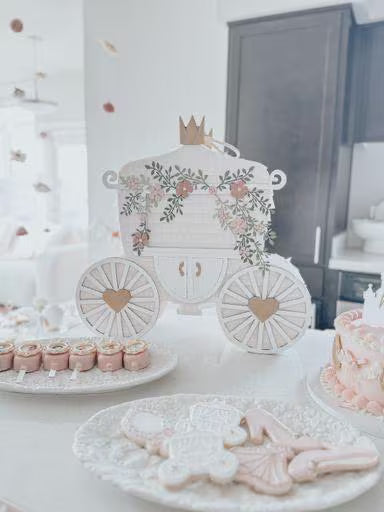 Regal carriage piñata with floral decorations and gold accents, placed on a table with pink desserts and cookies.