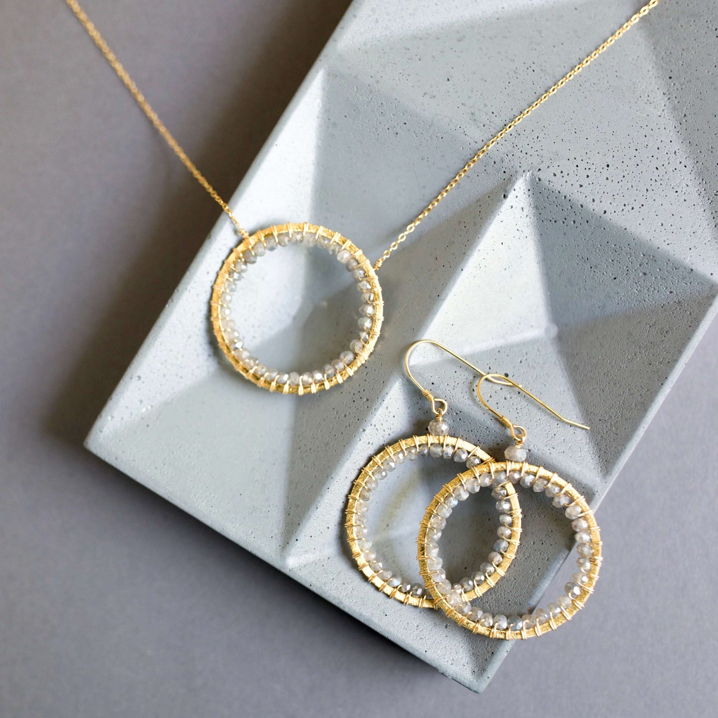 Gold wire-wrapped circular necklace and matching earrings with grey beads on a geometric stone tray.
