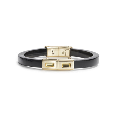 A black enamel bangle bracelet with gold-toned metal hardware, featuring a clasp engraved with 'JOY' and the 'ANNA S. LUX' brand logo, shown on a plain white background.