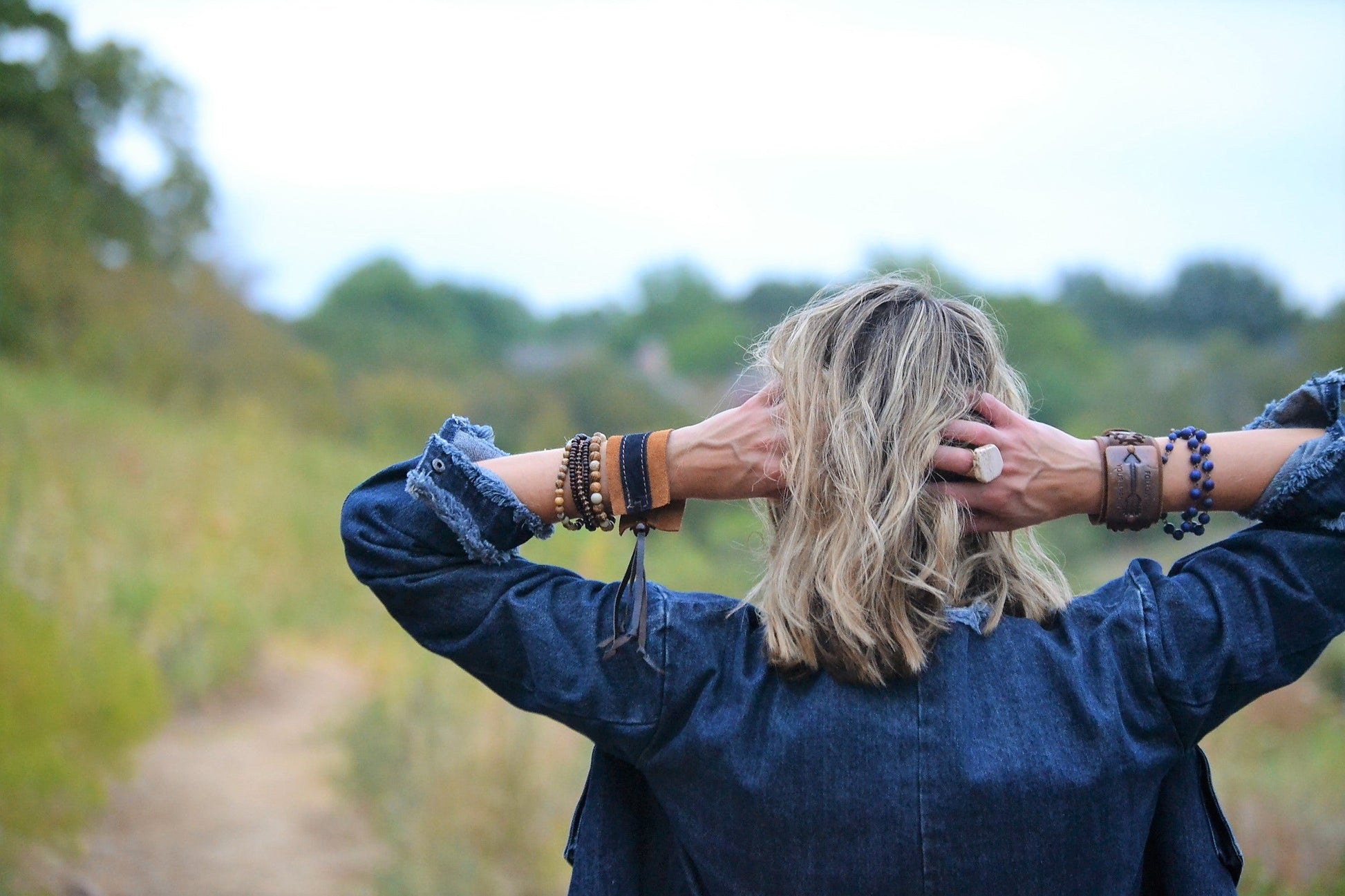 Person with blonde hair and multiple bracelets on arms, standing outdoors with blurred greenery in the background