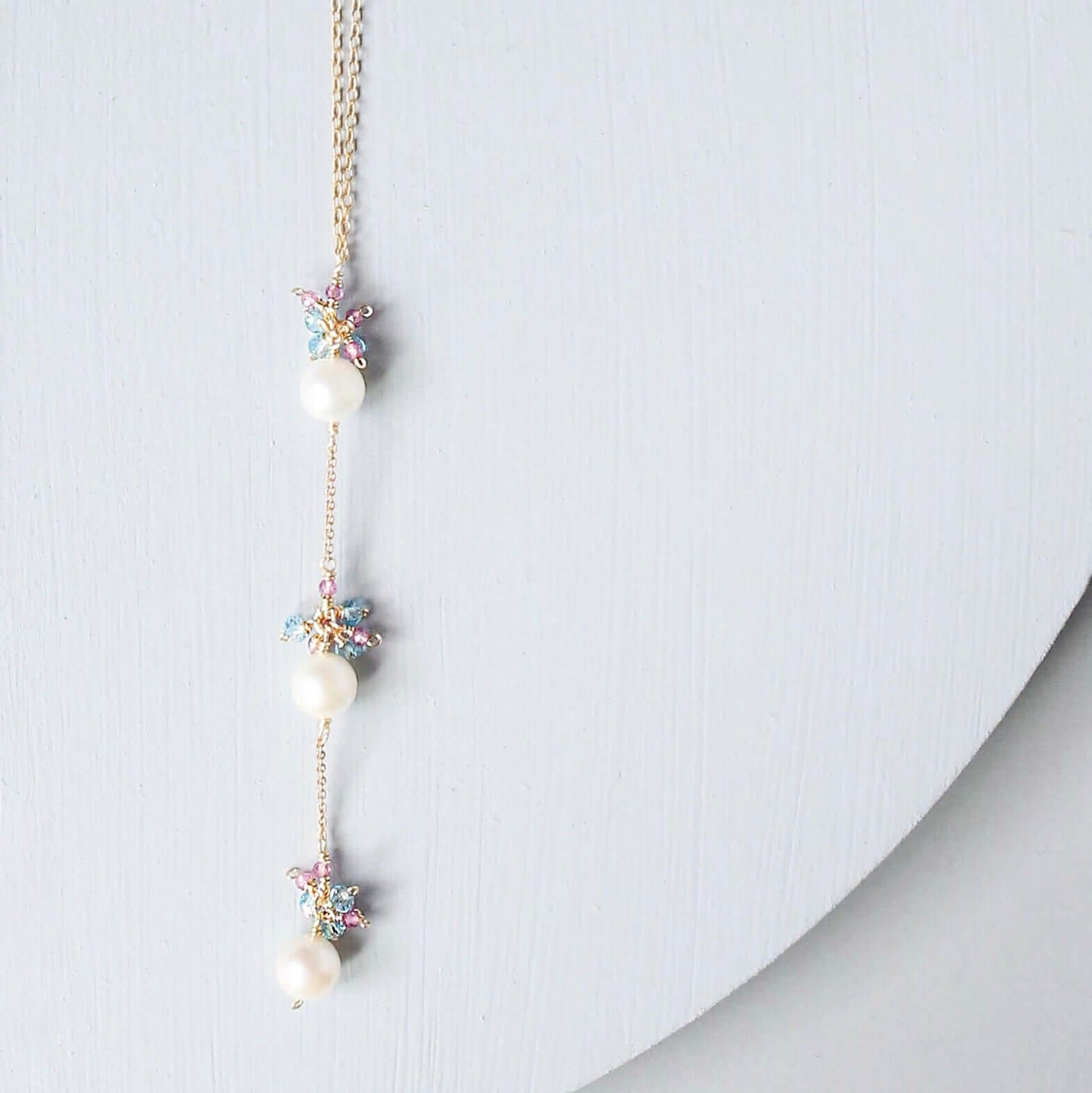 A delicate gold chain necklace featuring three white pearls and small pastel-colored starburst charms, displayed on a light gray curved surface.