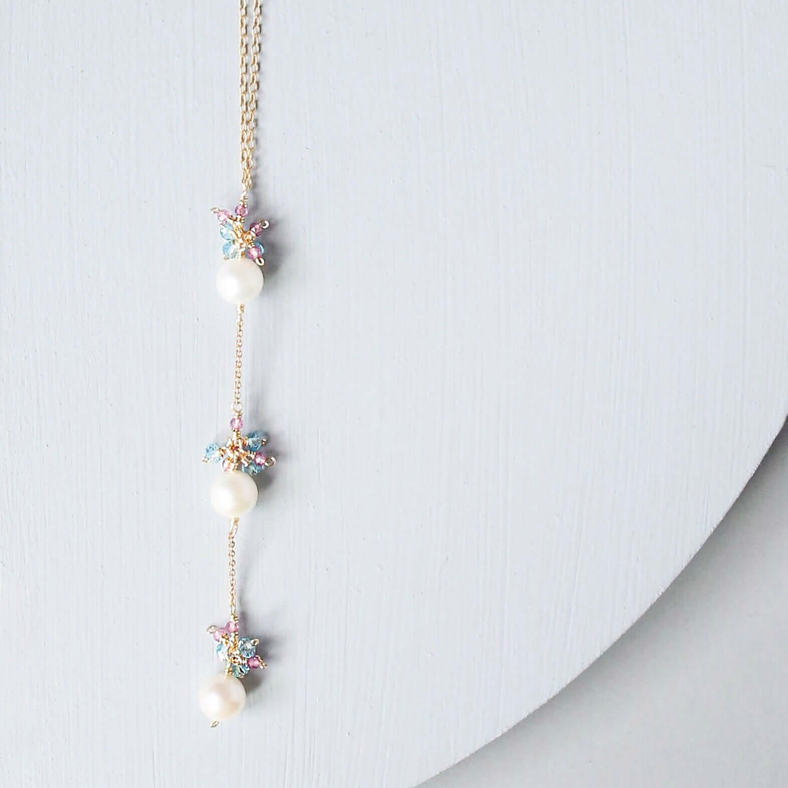 A delicate gold chain necklace featuring three white pearls and small pastel-colored starburst charms, displayed on a light gray curved surface.