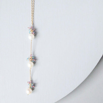 A delicate gold chain necklace featuring three white pearls and small pastel-colored starburst charms, displayed on a light gray curved surface.