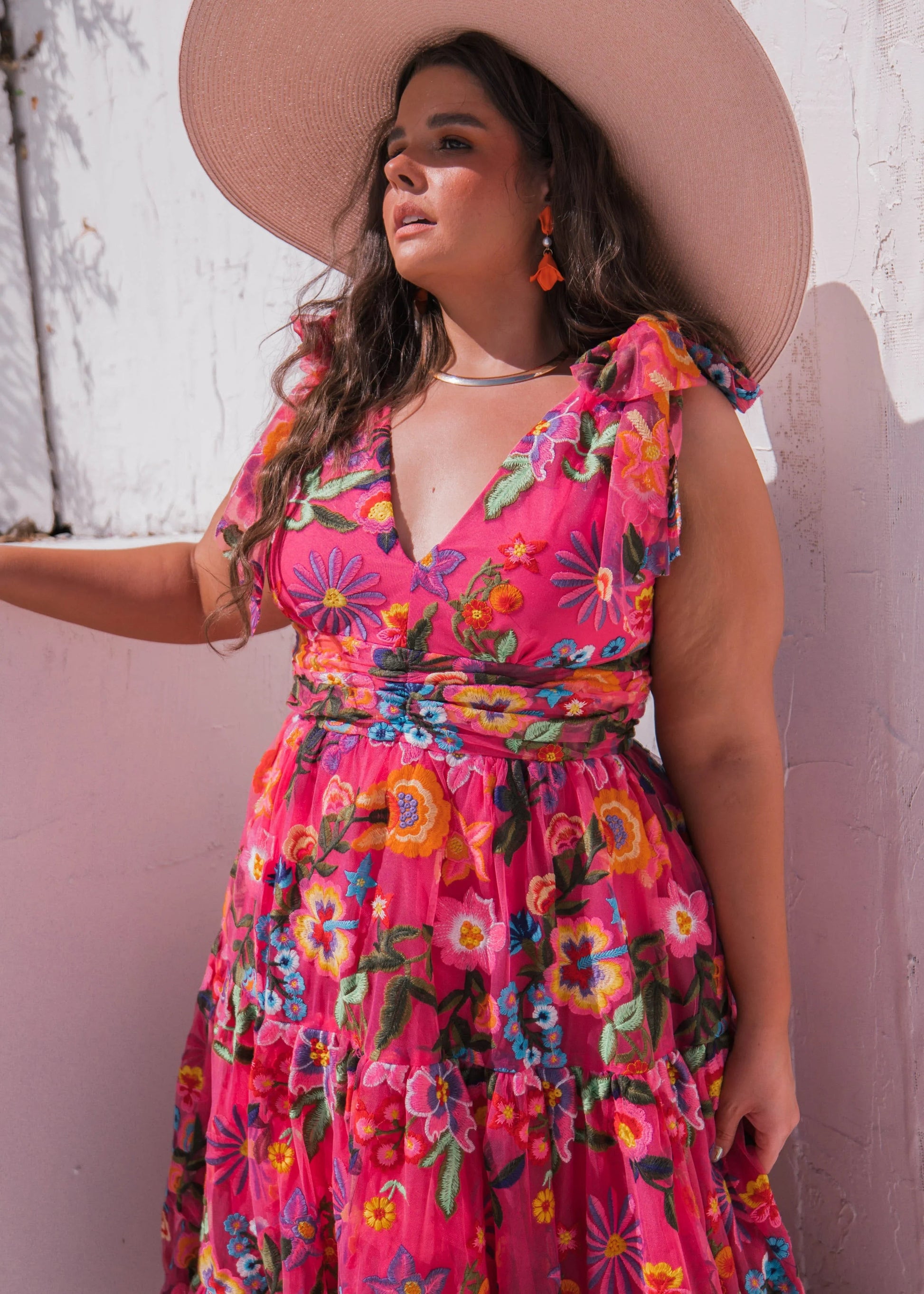 Woman wearing a colorful floral dress and wide-brimmed hat against a white wall.