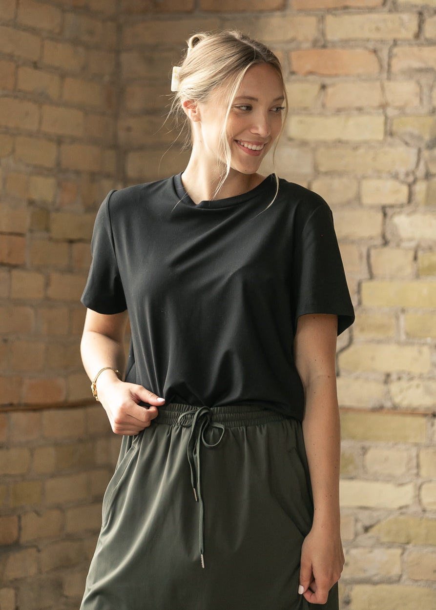 A woman wearing a black short-sleeve crewneck t-shirt and dark olive green drawstring pants, standing in front of a rustic brick wall.