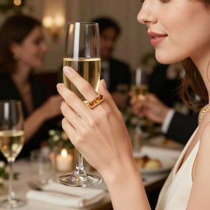 A young woman wearing an orange eternity ring holding a champagne glass with a group of people seated at a dinner table.