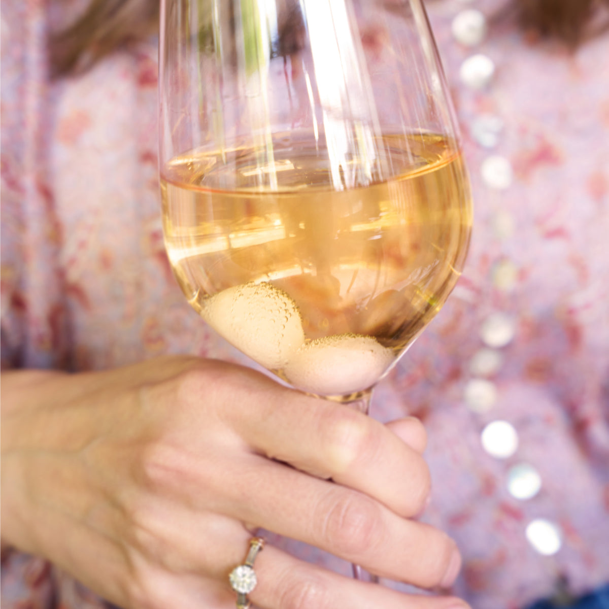 Hand holding a glass of white wine with rose quartz wine gems at the bottom.