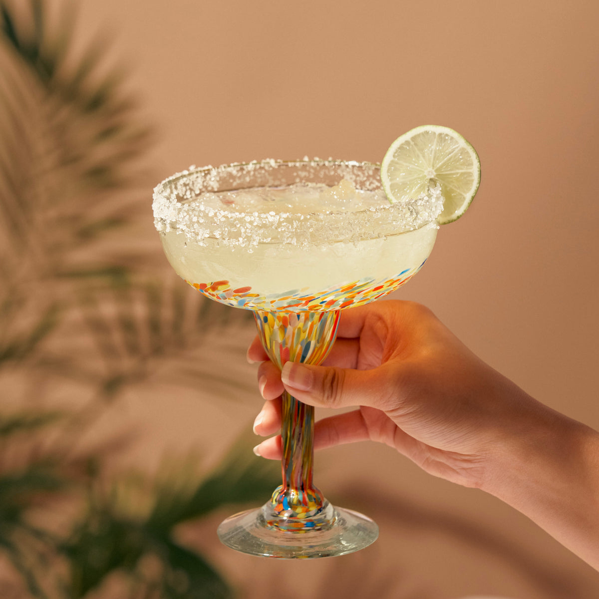 Hand holding a colorful speckled margarita glass filled with a drink, with a salted rim and lime garnish.