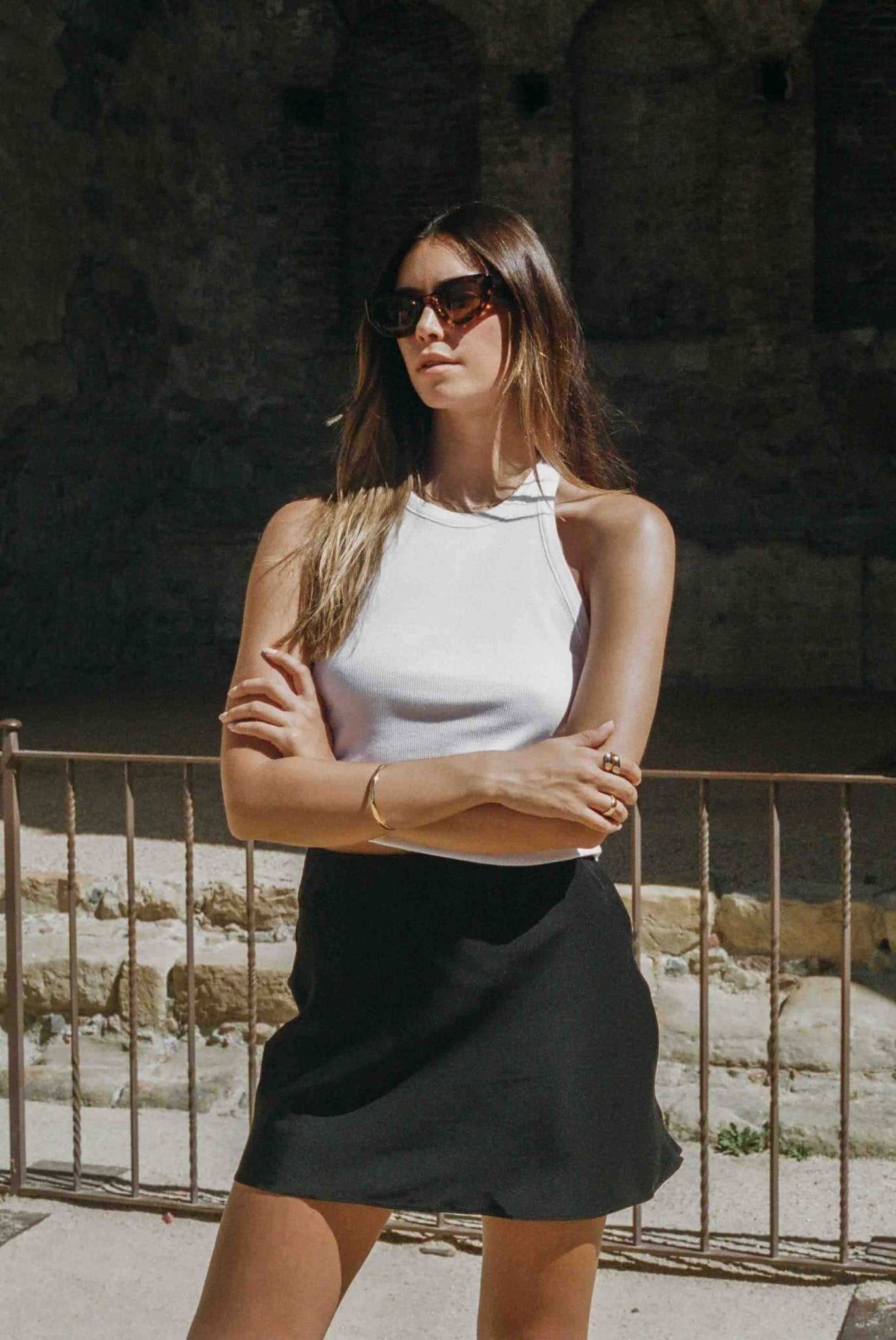 Woman wearing sunglasses and a white top with a black skirt standing in front of stone walls.