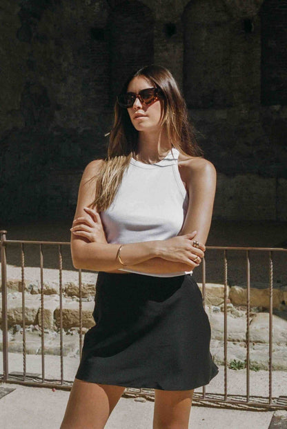 Woman wearing sunglasses and a white top with a black skirt standing in front of stone walls.