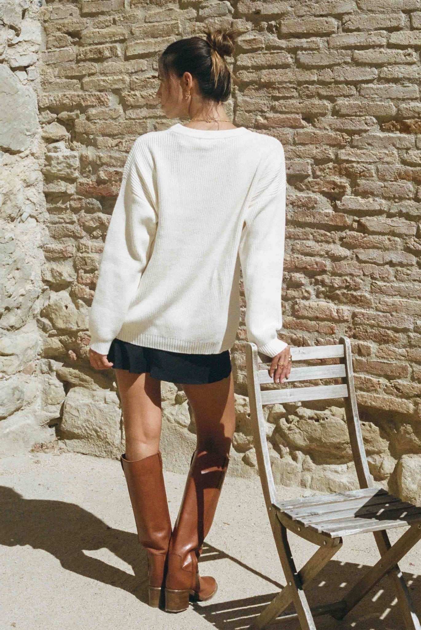 Person wearing a white sweater and brown knee-high boots standing against a stone wall.