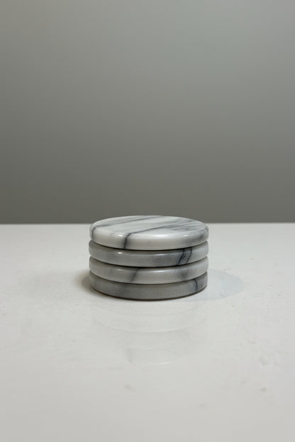 Four round marble coasters with a white and gray veined pattern, arranged in a row on a light gray textured surface.