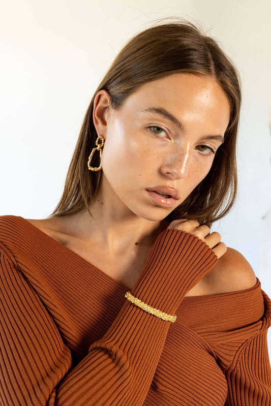 A woman wearing a textured gold Nicole Cuff Bracelet over the sleeve of a brown ribbed knit top.