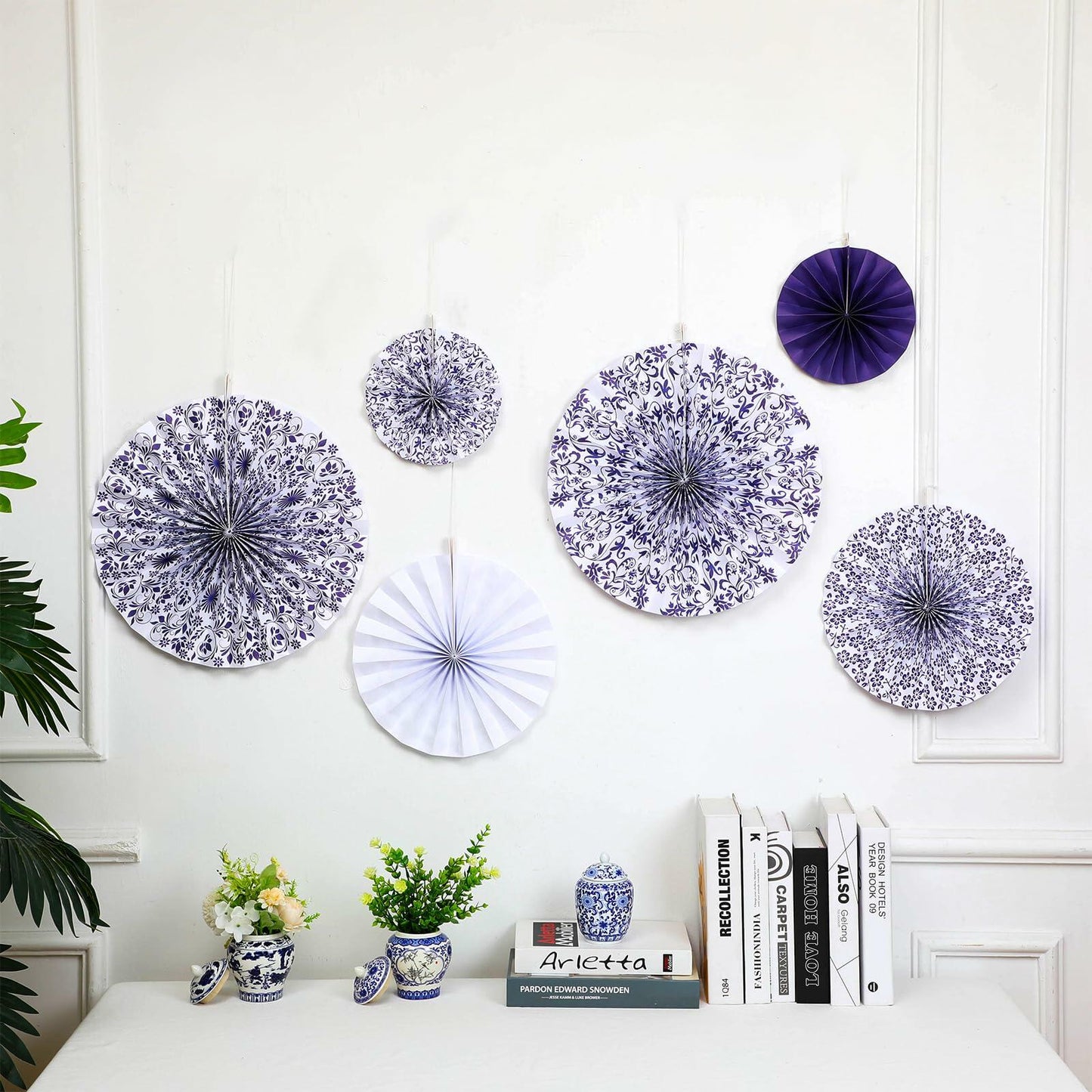 Chinoiserie hanging paper fans in blue and white, featuring floral patterns, arranged on a wall with decorative items below.